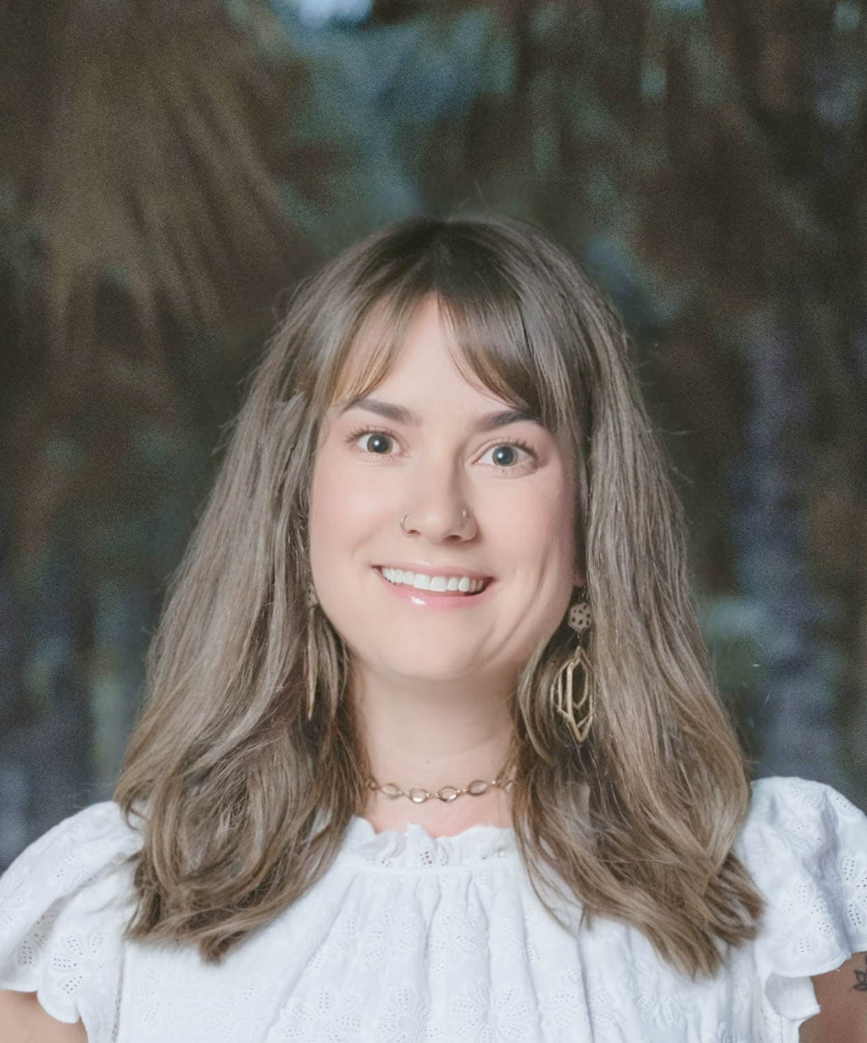 Kristin-Mills-01 Woman with shoulder-length brown hair, in a white eyelet blouse and gold earrings, smiles at the camera outdoors with blurred trees in the background.