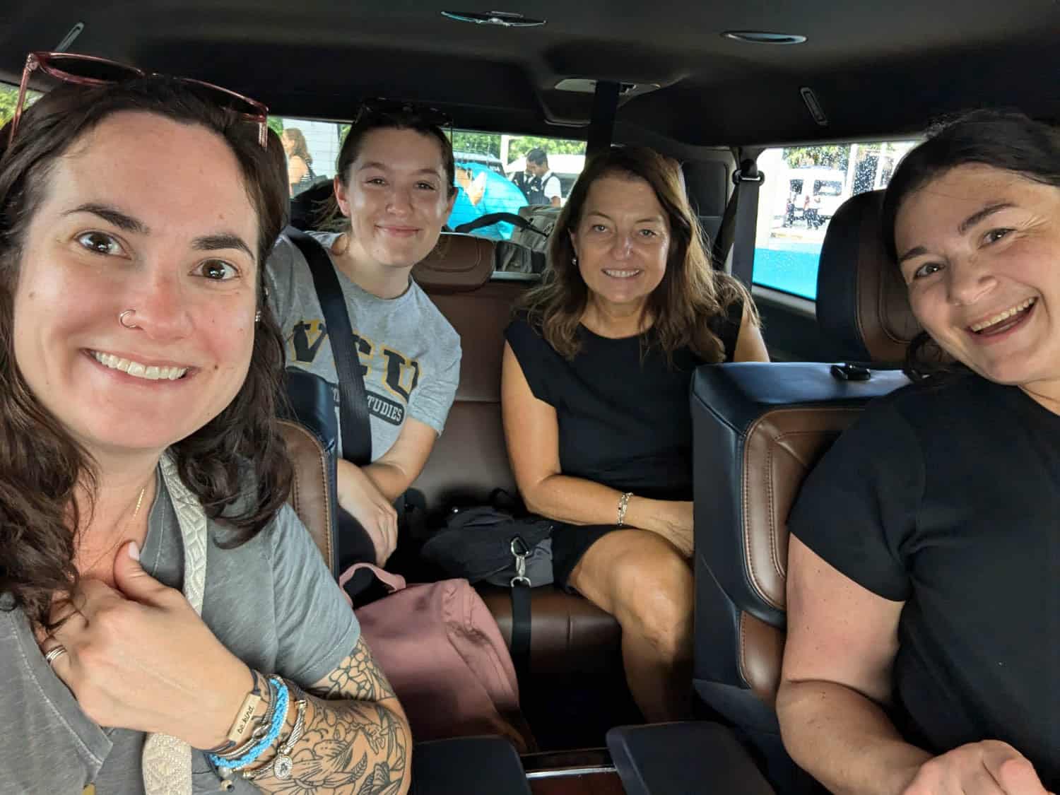 Kristin-Mills-04 Four women sit close together inside a vehicle, smiling at the camera. One has a tattooed arm and wears a nose ring. Bags are visible on the seats.