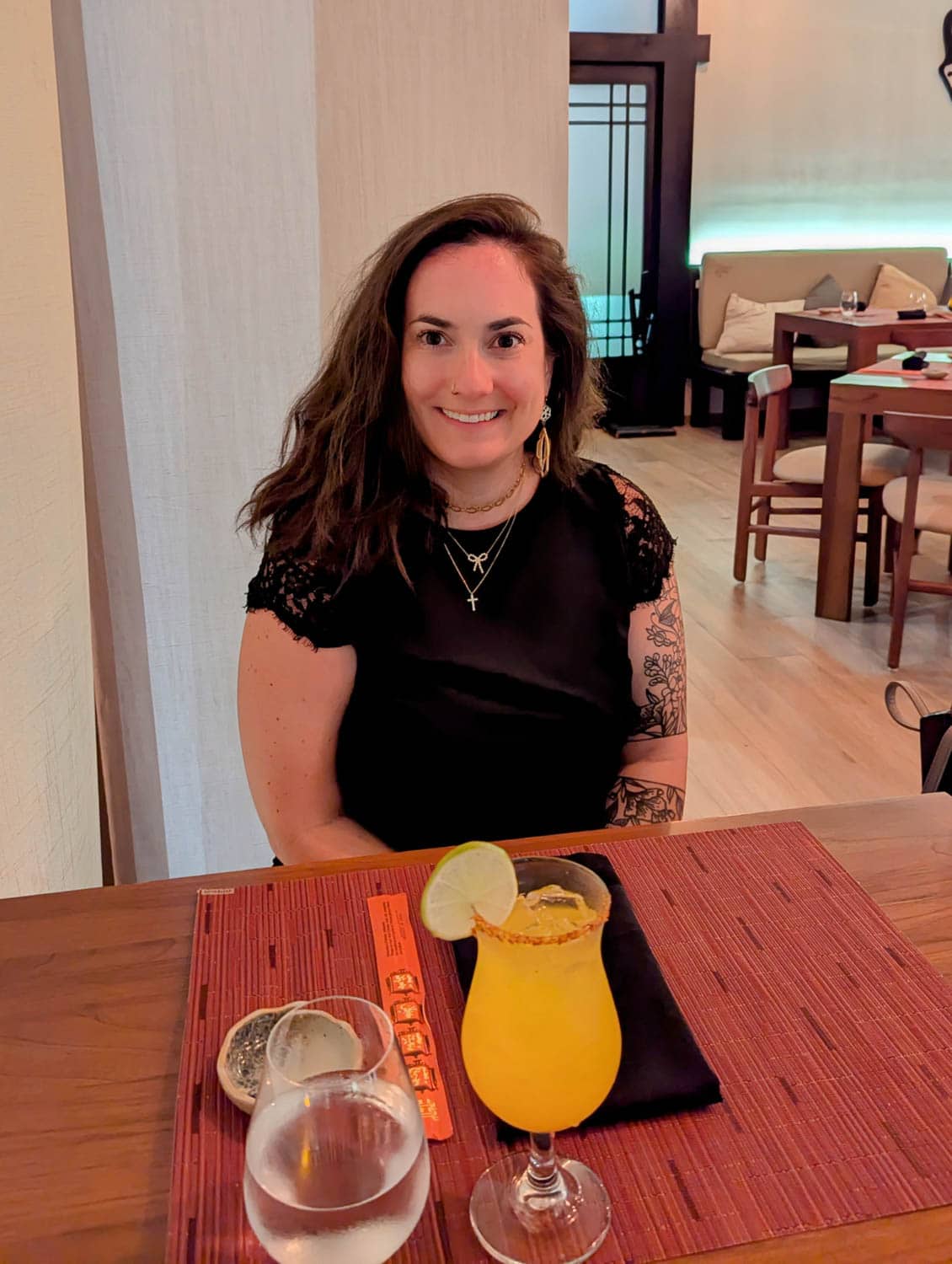 Kristin-Mills-05 A woman with long brown hair and a black dress sits at a table with a yellow cocktail, a glass of water, and chopsticks in a modern restaurant.