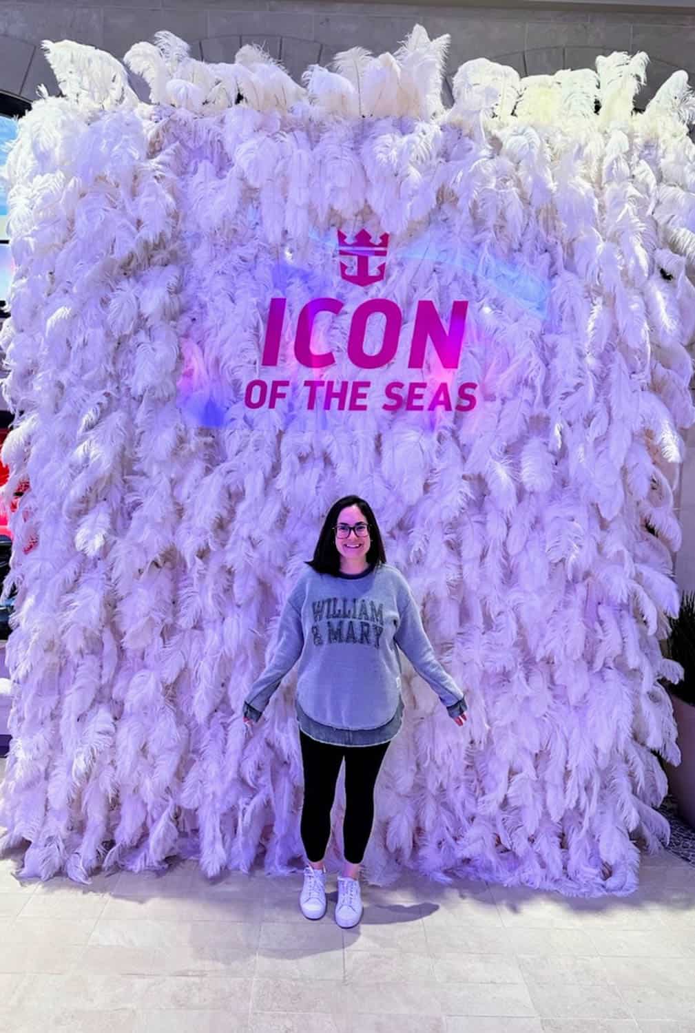 Kristin-Mills-09 A woman stands in front of a white feather backdrop with the text ICON OF THE SEAS in pink and purple lettering above them.