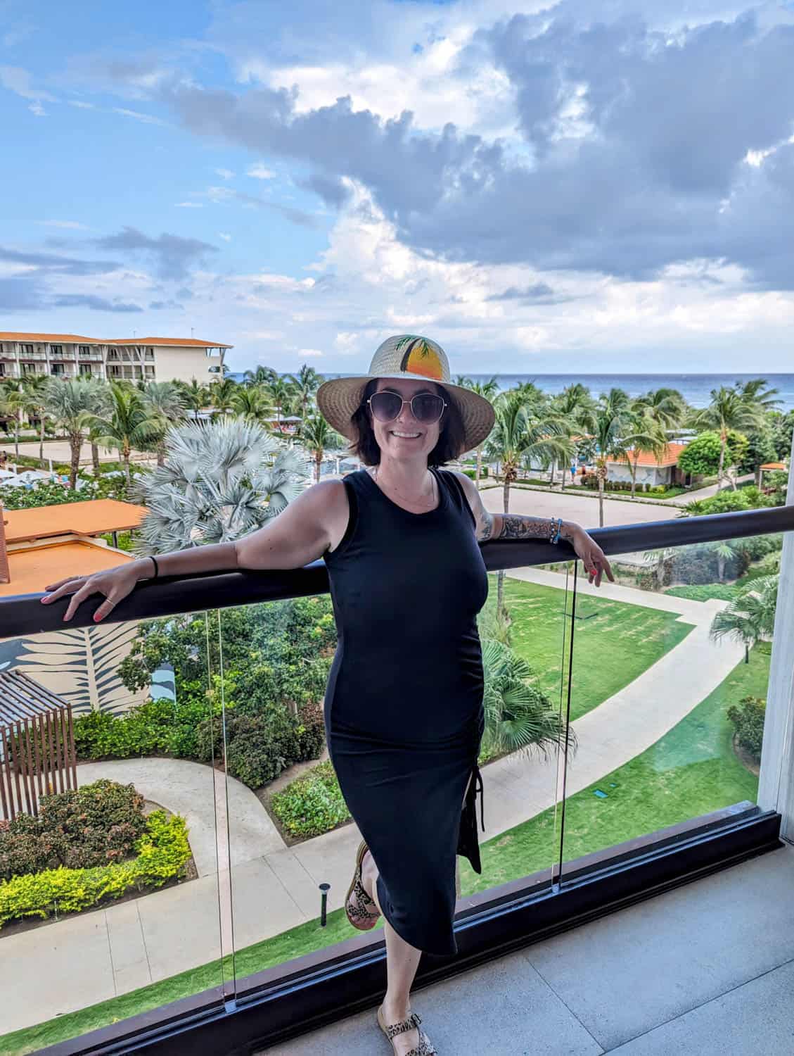 Kristin-Mills-10 A woman in a black dress and sun hat stands on a balcony overlooking a resort with palm trees, gardens, and an ocean view in the background.
