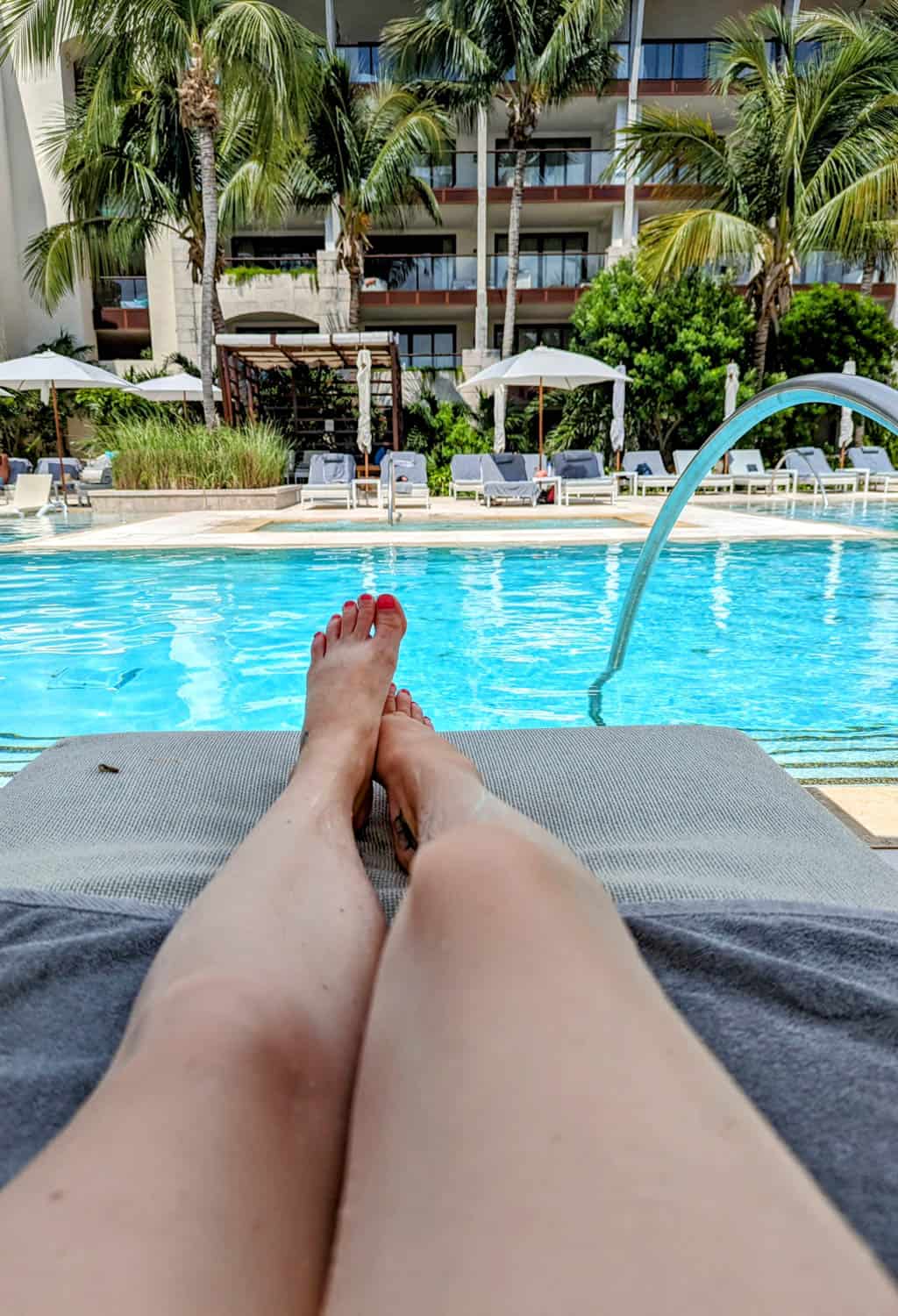 Kristin-Mills-11 Woman lounging on a chair by a swimming pool with palm trees, umbrellas, and a hotel building in the background.