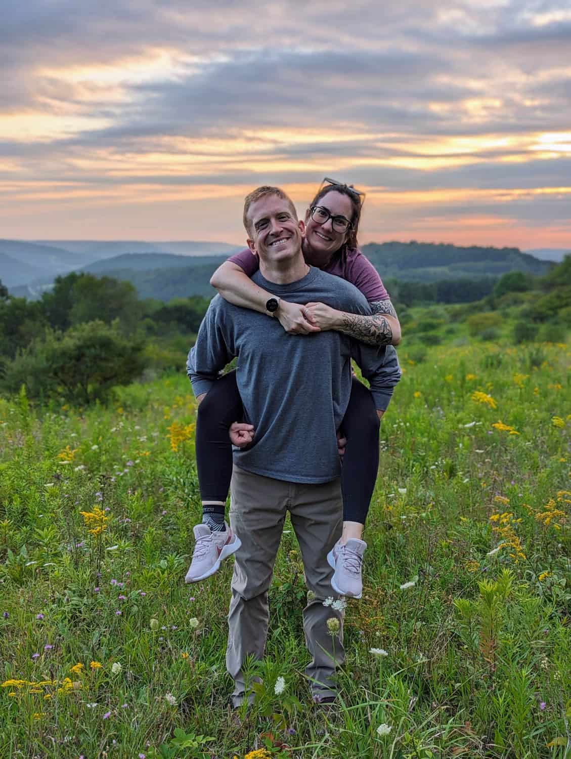 Kristin-Mills-12 A man stands in a wildflower field at sunset with a woman on his back, both smiling at the camera. Rolling hills and cloudy sky are visible in the background.