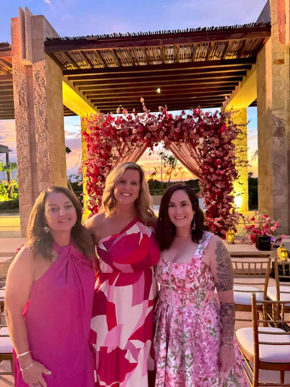 Kristin-Mills-13 Three women in pink dresses stand in front of a floral arch at an outdoor event during sunset.