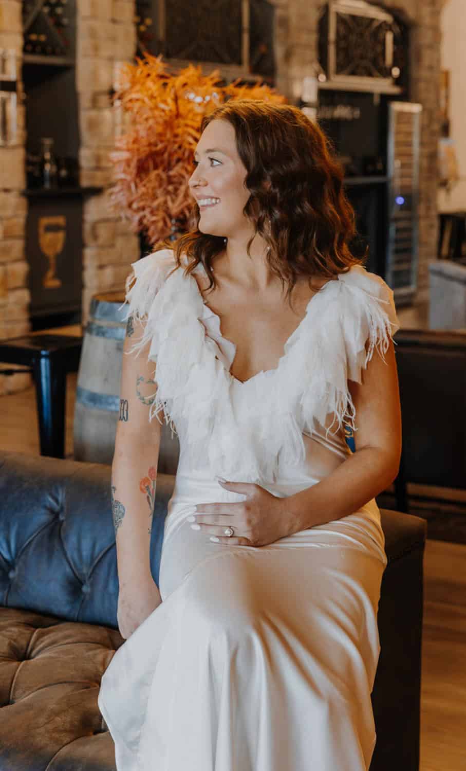 Madelynn-Pierce-01 A woman in a white dress with feather details sits on a dark couch, smiling and looking to the side in a warmly lit room.