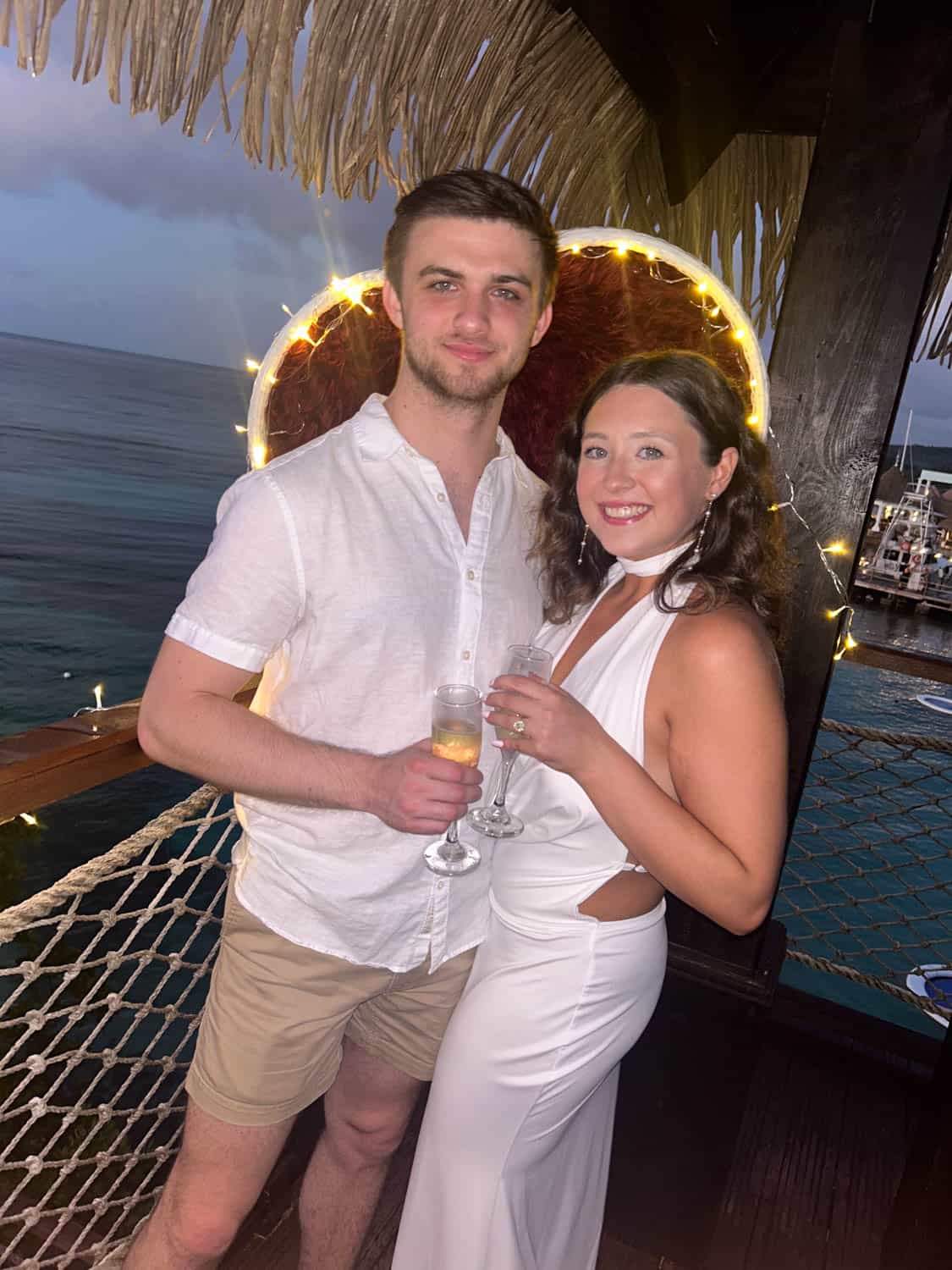 Madelynn-Pierce-04 A man and woman stand together on a deck over the ocean, dressed in white, holding drinks and smiling at the camera with string lights in the background.