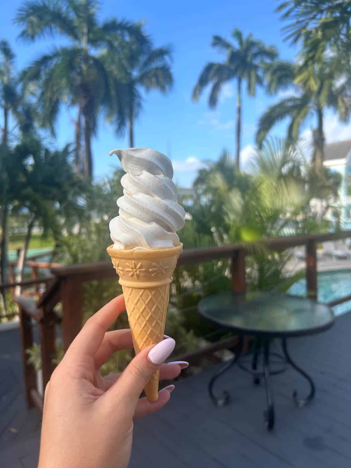 Madelynn-Pierce-05 A hand holding a vanilla soft serve ice cream cone outdoors, with palm trees and a round table in the background.