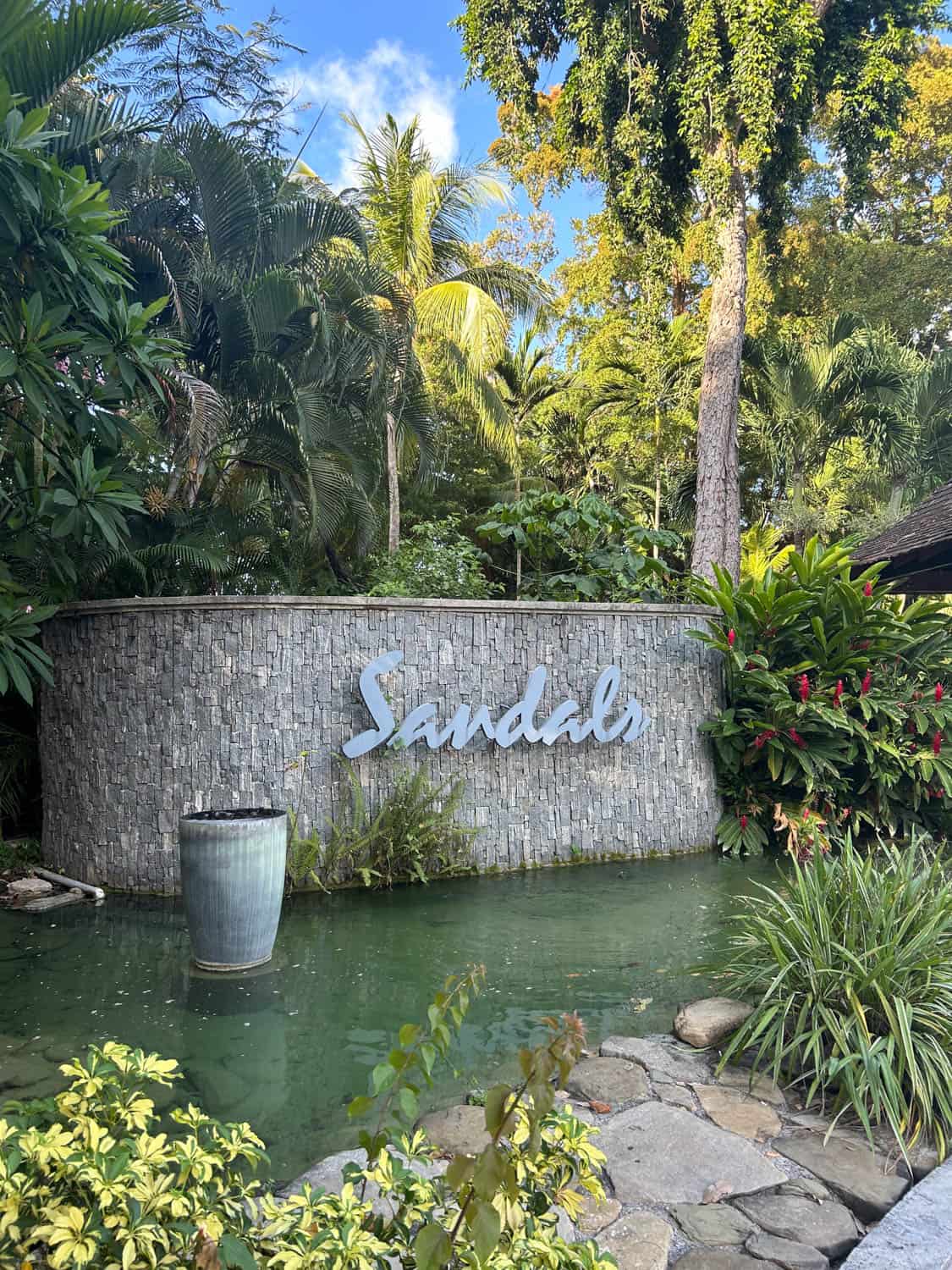 Madelynn-Pierce-07 A stone wall with the word Sandals in large letters stands behind a small pond and lush tropical plants.