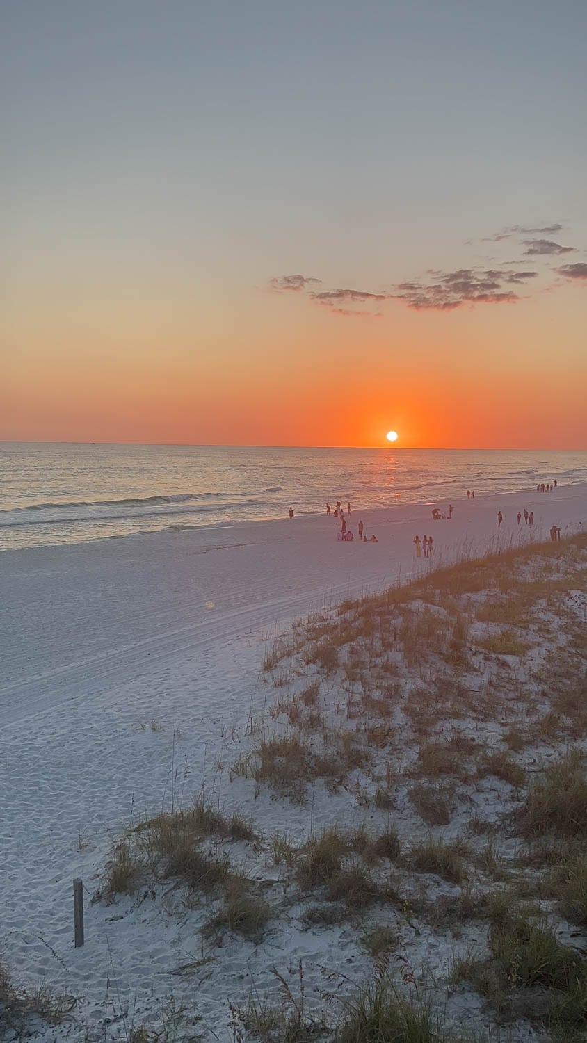 Madelynn-Pierce-09 Sunset over a sandy beach with people walking and gathering near the shoreline. Grassy dunes are visible in the foreground.