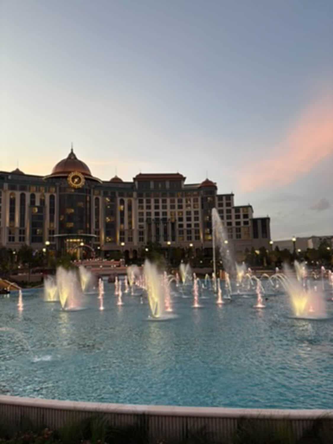 Madelynn-Pierce-11 Large hotel building with a dome and clock faces a fountain with illuminated water jets at dusk.