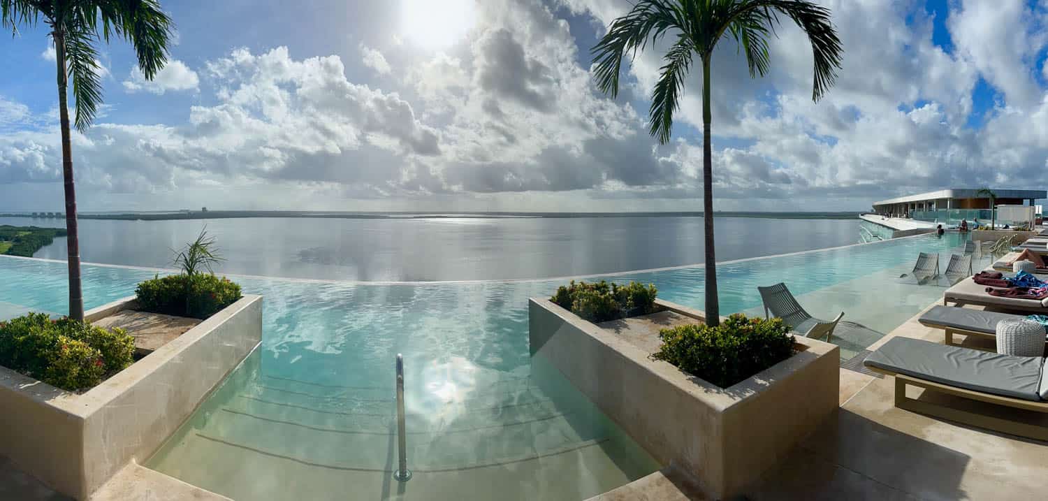 Infinity pool overlooking a calm body of water with palm trees, lounge chairs, and people relaxing under a partly cloudy sky in Mexico.