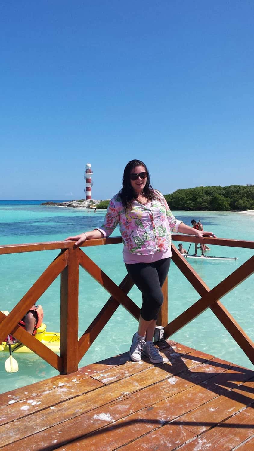 A woman stands on a wooden deck by clear turquoise water with a red-and-white lighthouse and small groups of people in the background under a clear blue sky.