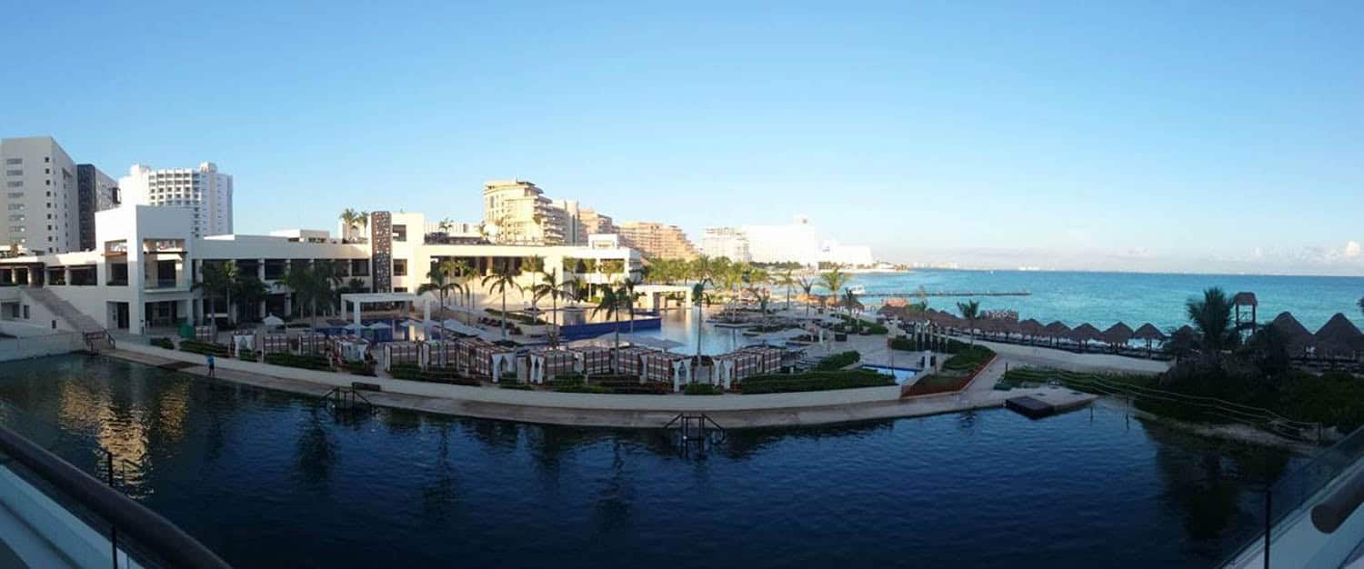 View of a beachfront resort with pools, lounge chairs, palm trees, and buildings in the background, overlooking the ocean under a clear blue sky.