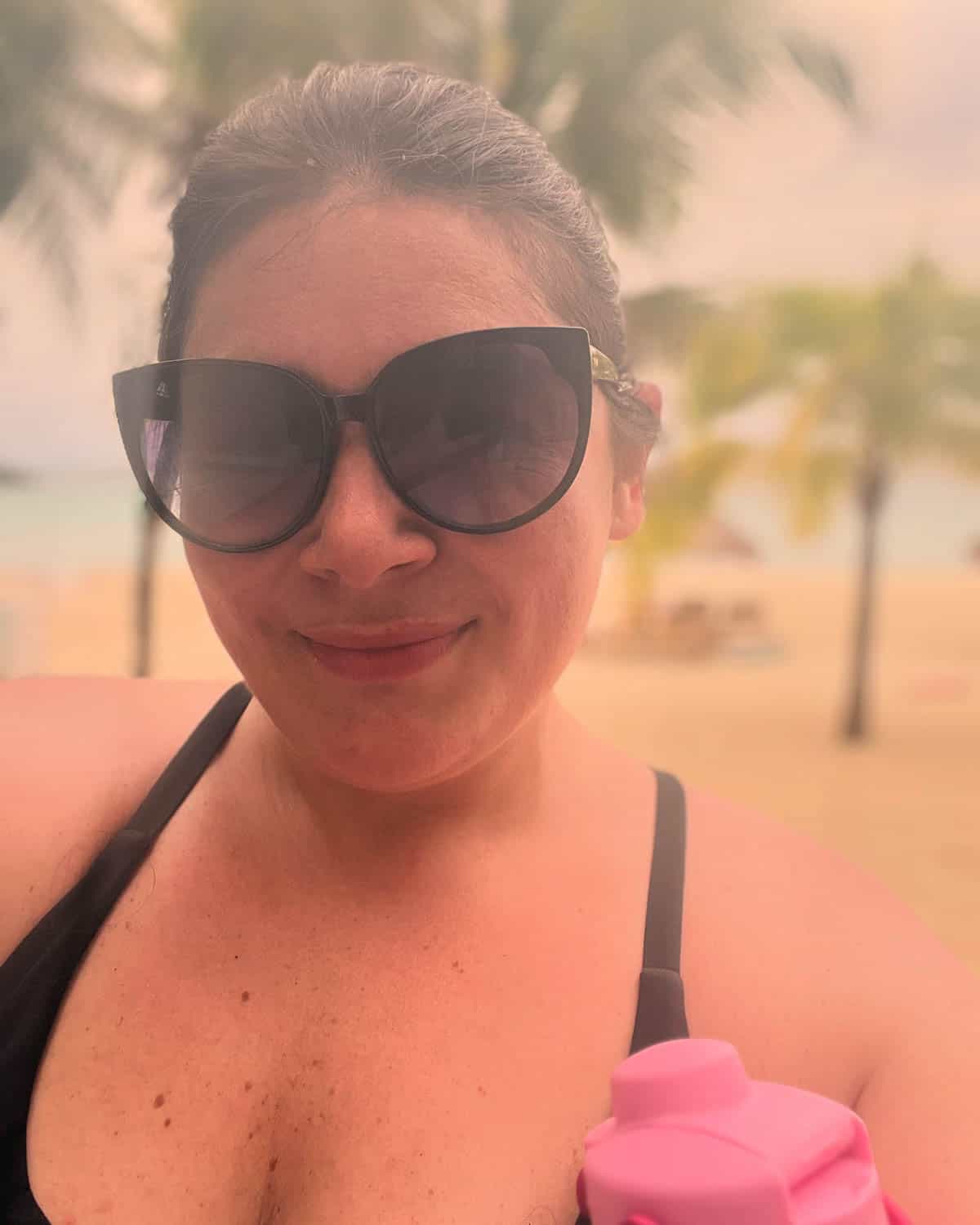 Woman wearing large black sunglasses and a swimsuit, holding a pink water bottle, with a beach and palm trees in the background.