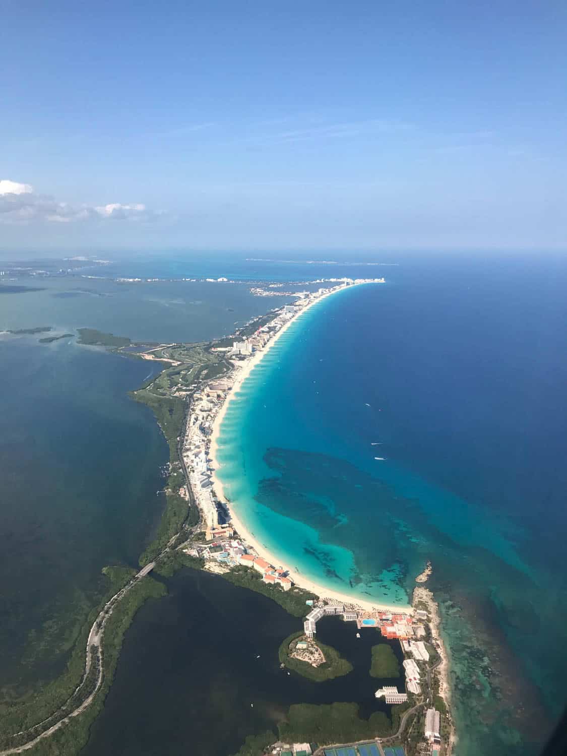 Aerial view of a curved coastline with white sand beaches, turquoise water, city buildings, deep blue ocean, and surrounding green landscape.