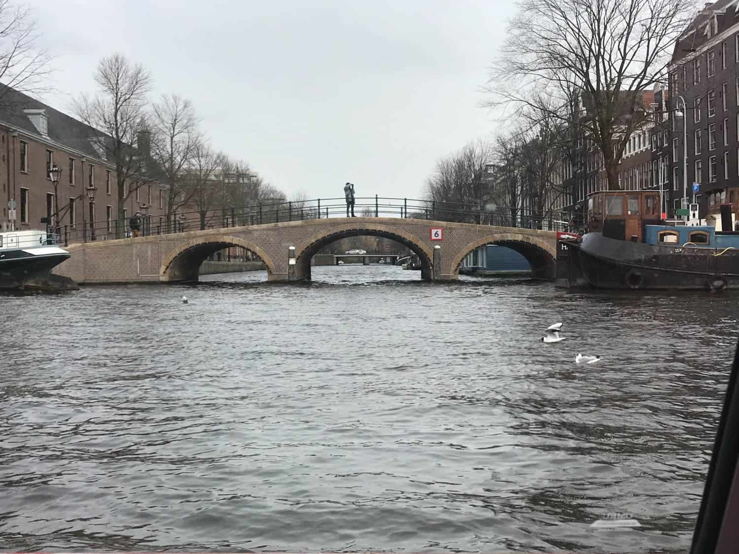 A stone bridge with three arches spans a canal lined with boats and buildings on a cloudy day. A person stands on the bridge and birds float on the water.