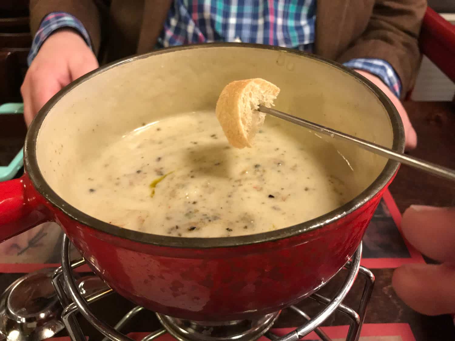 A piece of bread on a fork is being dipped into a pot of cheese fondue, with a person in a plaid shirt and brown jacket seated in the background.