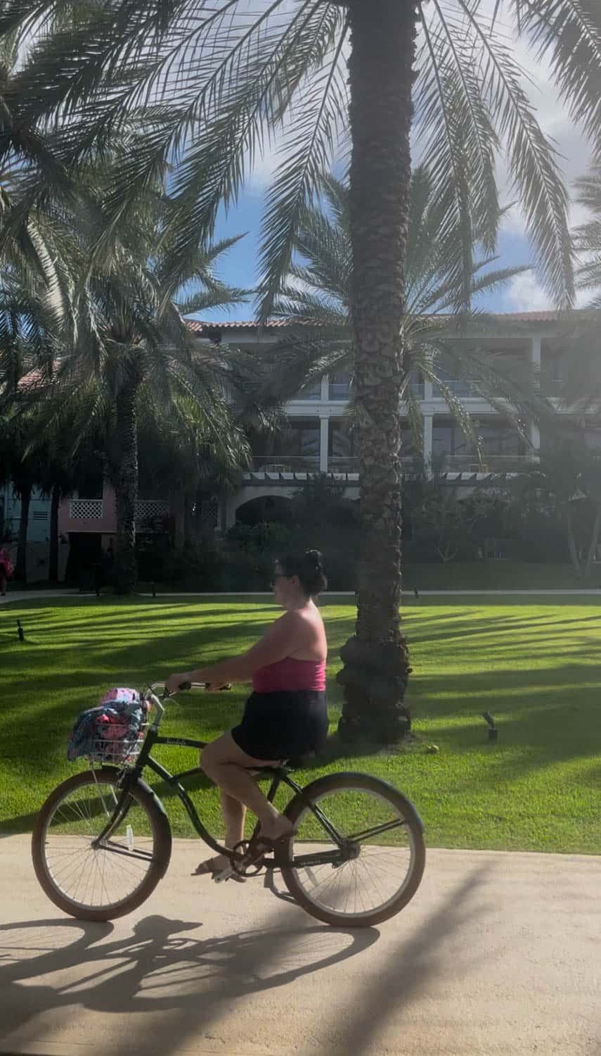 A woman rides a bicycle on a paved path through a sunlit lawn with palm trees and a large building in the background.