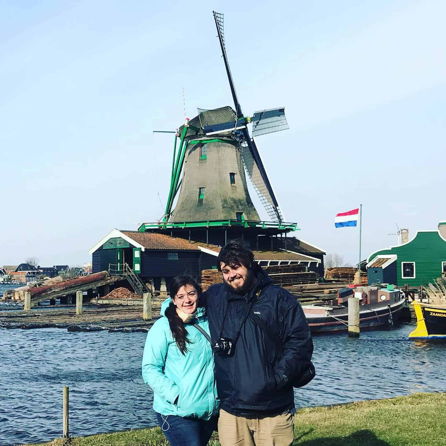 Two people stand smiling by the water with a traditional Dutch windmill and the Netherlands flag in the background.