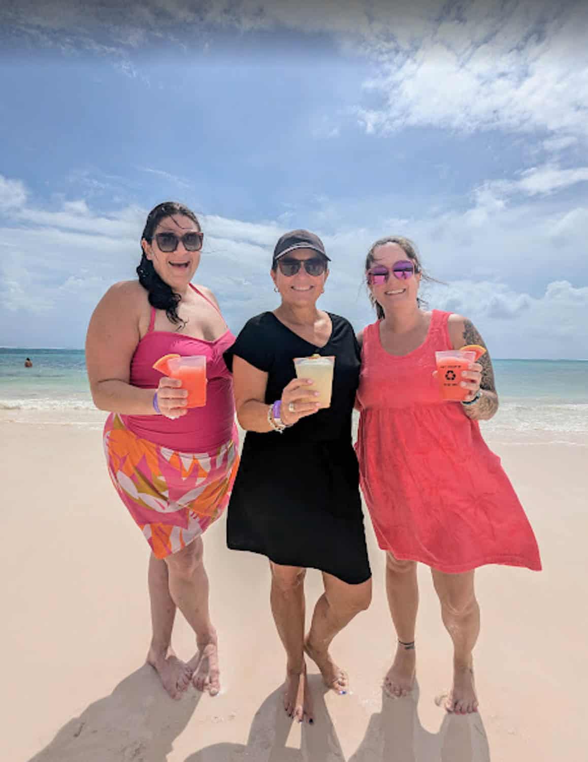 Three women stand barefoot on a sandy beach, each holding a drink, with the ocean and a partly cloudy sky in the background.