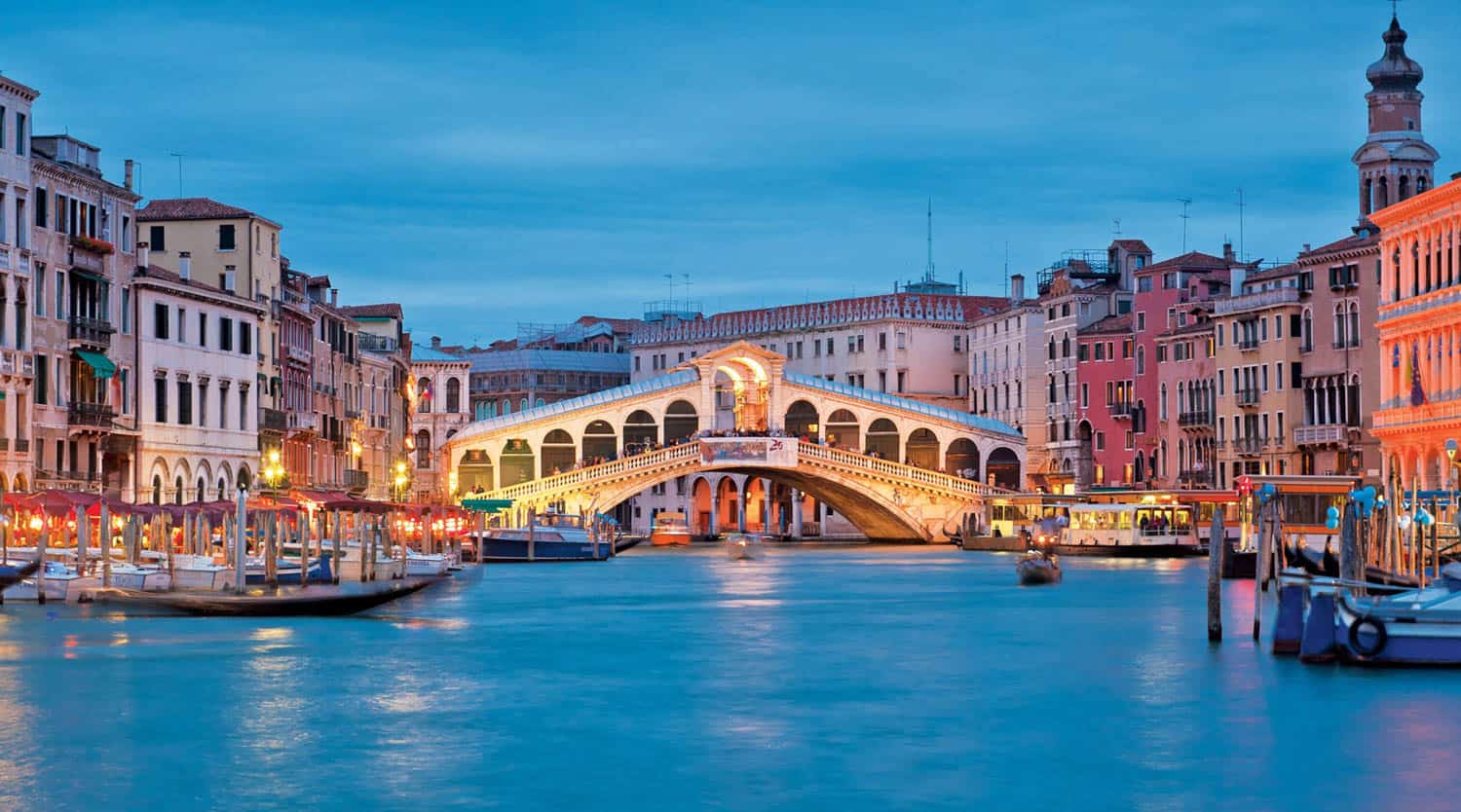 Venice's Grand Canal with the illuminated Rialto Bridge at dusk, surrounded by historic buildings and boats along the waterfront.