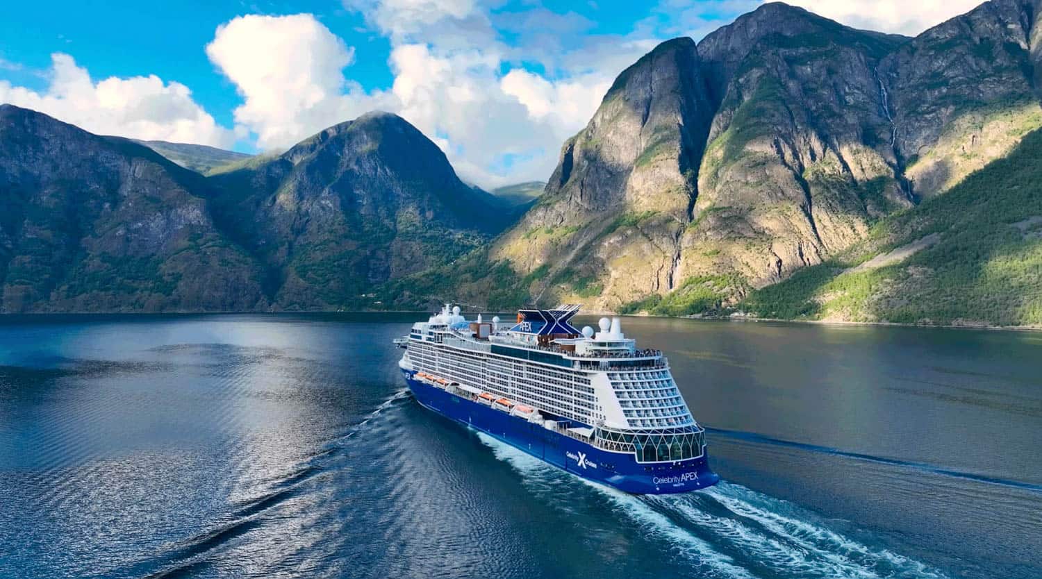 A large blue and white cruise ship sails through calm water surrounded by tall, green mountains under a partly cloudy sky.