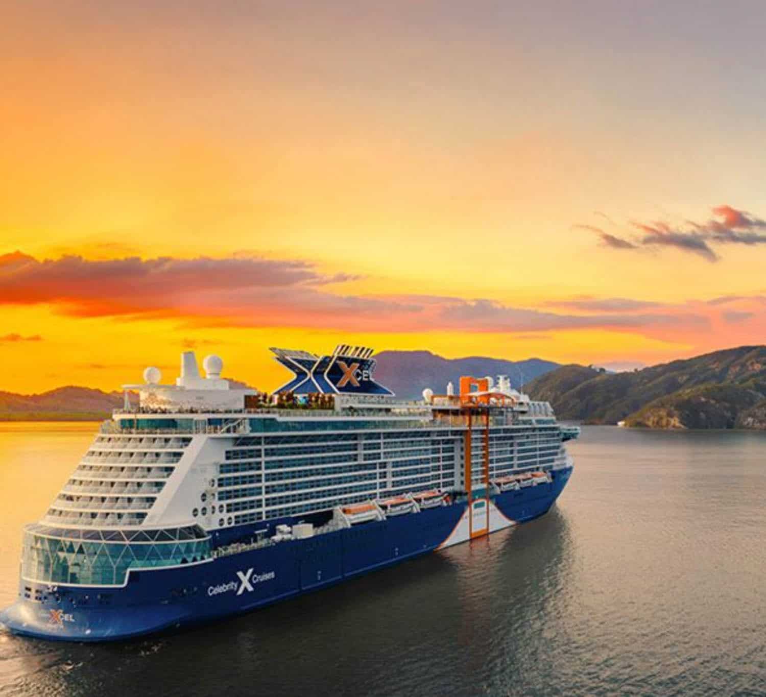 A large blue and white cruise ship sails near a coastline at sunset, with mountains and an orange sky in the background.