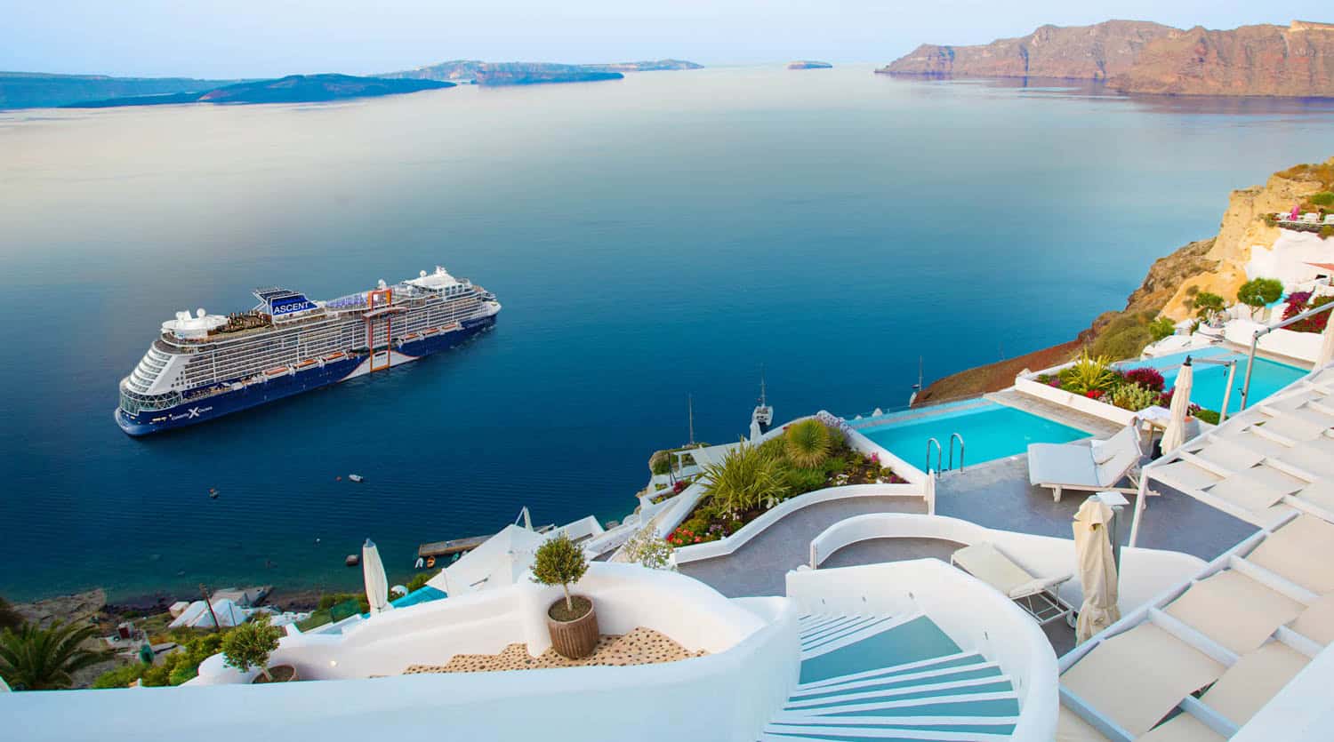 A large cruise ship is anchored in a calm blue bay near steep coastal cliffs with white buildings and terraced pools overlooking the water.