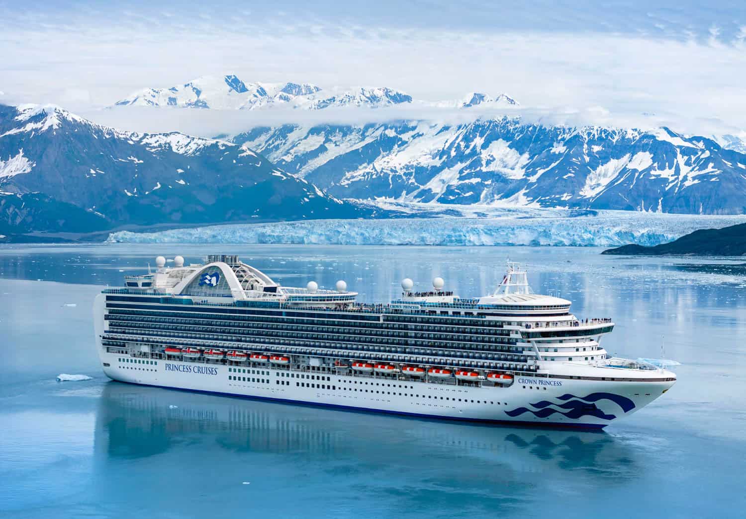 A large Princess Cruises ship sails in calm blue water with snow-capped mountains and a glacier in the background in Alaska.