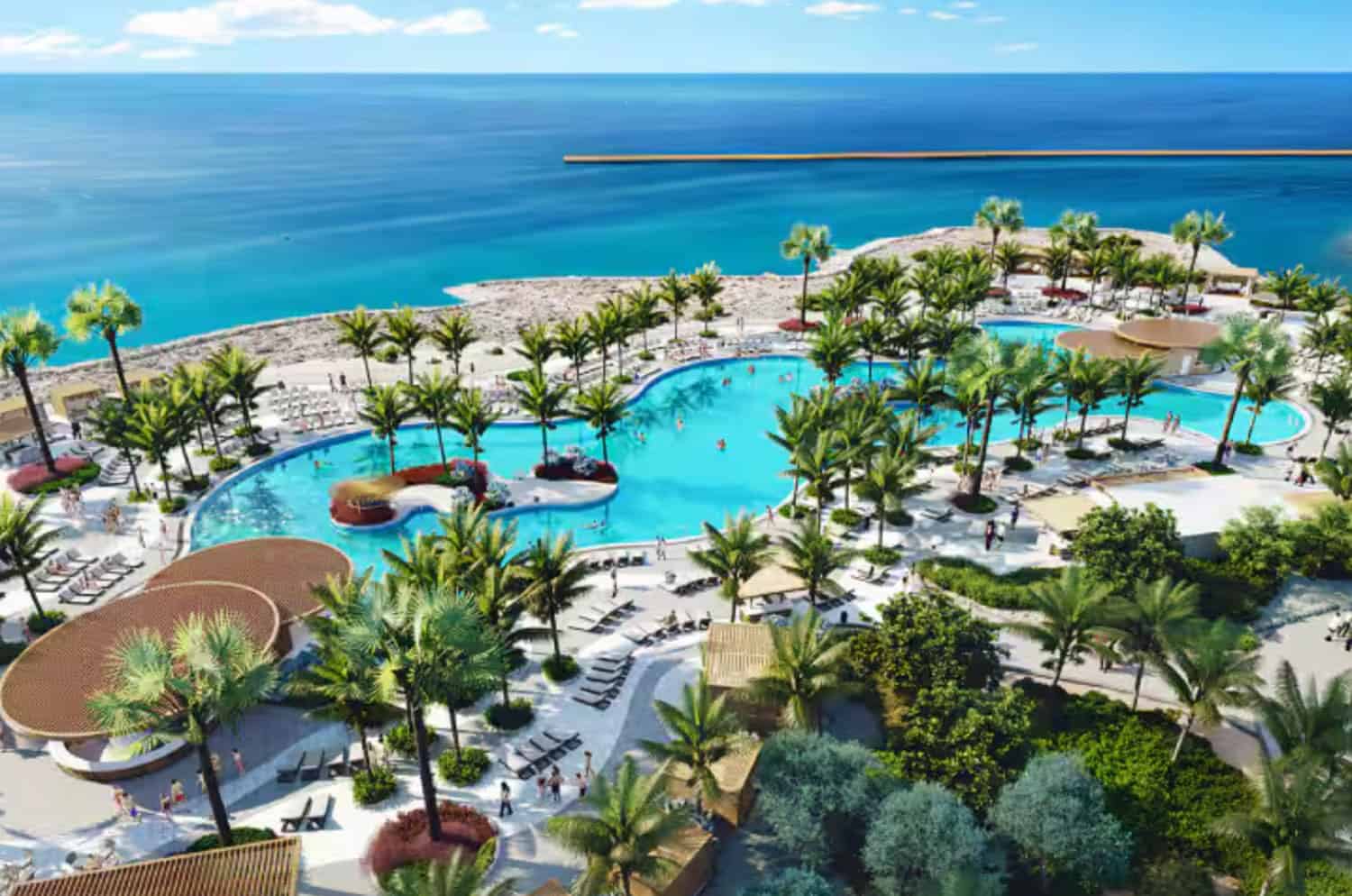 Aerial view of a large resort pool surrounded by palm trees, lounge chairs, and landscaped gardens, with the ocean and a rocky pier in the background.