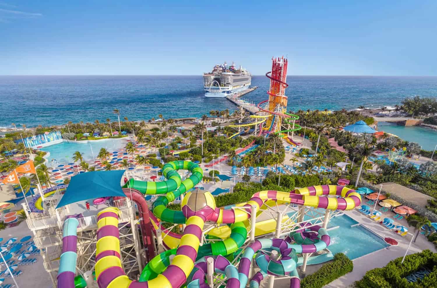 Aerial view of a colorful water park with winding slides near the ocean, with a cruise ship docked in the background.