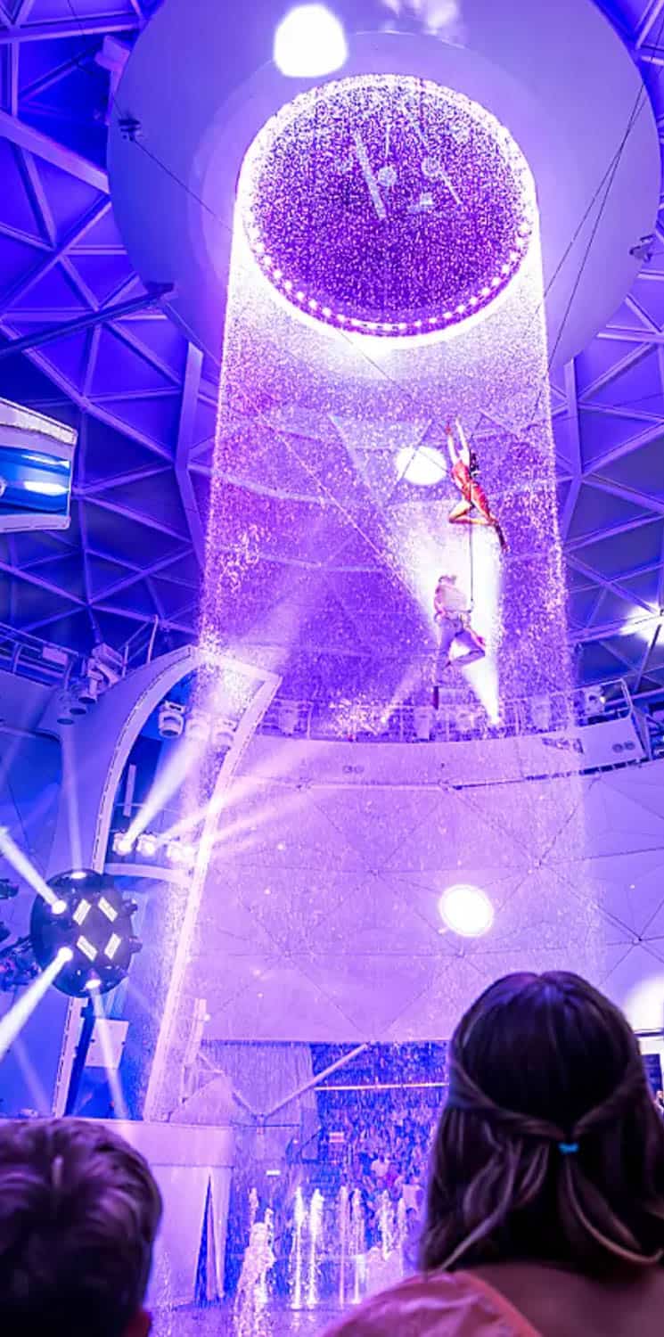 Acrobats perform on suspended apparatus under a circular light feature with falling water, while an audience watches from below in an indoor venue on a cruise.