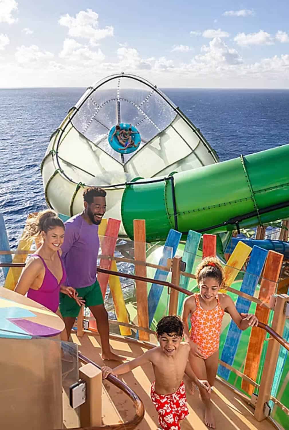A family of four stands at the entrance of a large green waterslide on a cruise ship, with an ocean view in the background.