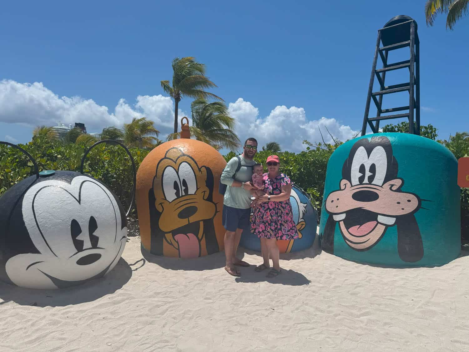 Olivia-Williams-01 Two adults and a baby on a beach in front of large painted buoys featuring Mickey Mouse, Pluto, and Goofy faces. Palm trees and blue sky are in the background.