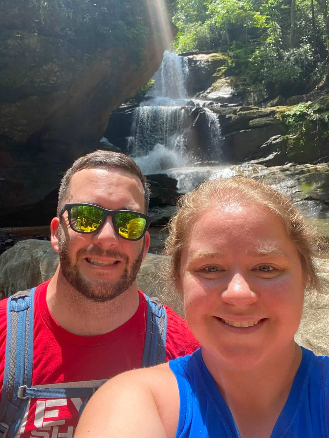 Olivia-Williams-02 Two people smiling for a selfie in front of a waterfall surrounded by rocks and greenery on a sunny day.