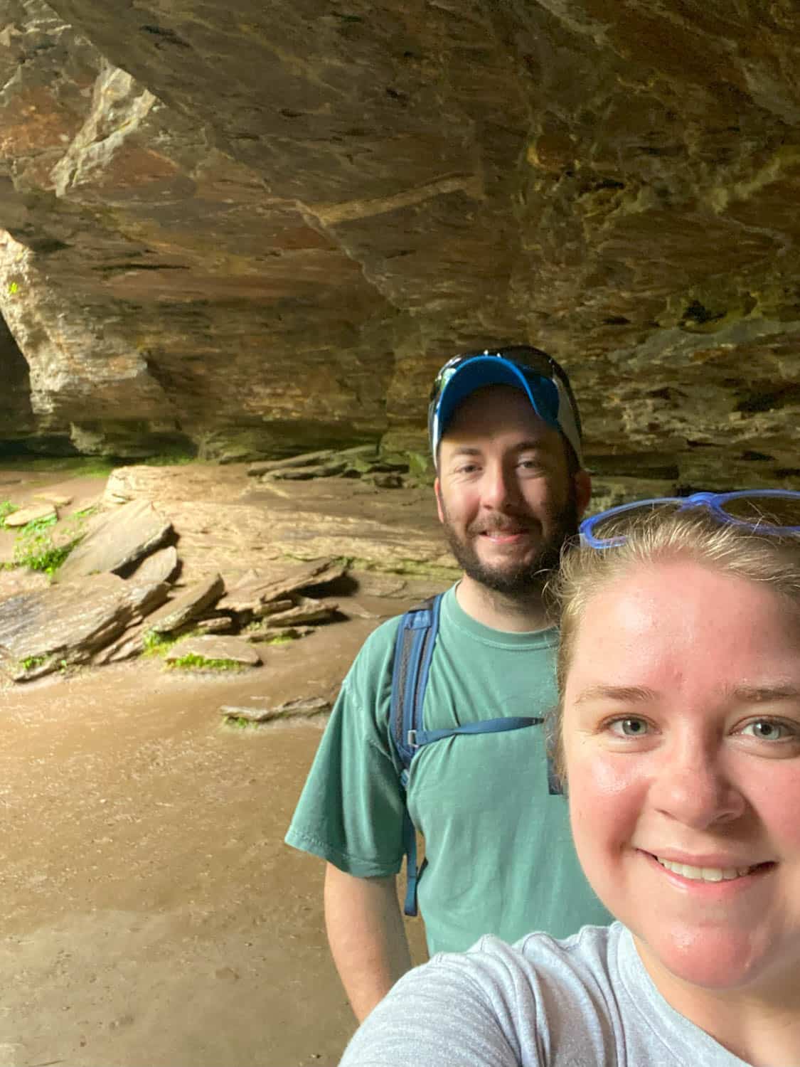 Olivia-Williams-04 Two people smiling for a selfie inside a rocky cave or cavern with uneven rock surfaces and some greenery in the background.