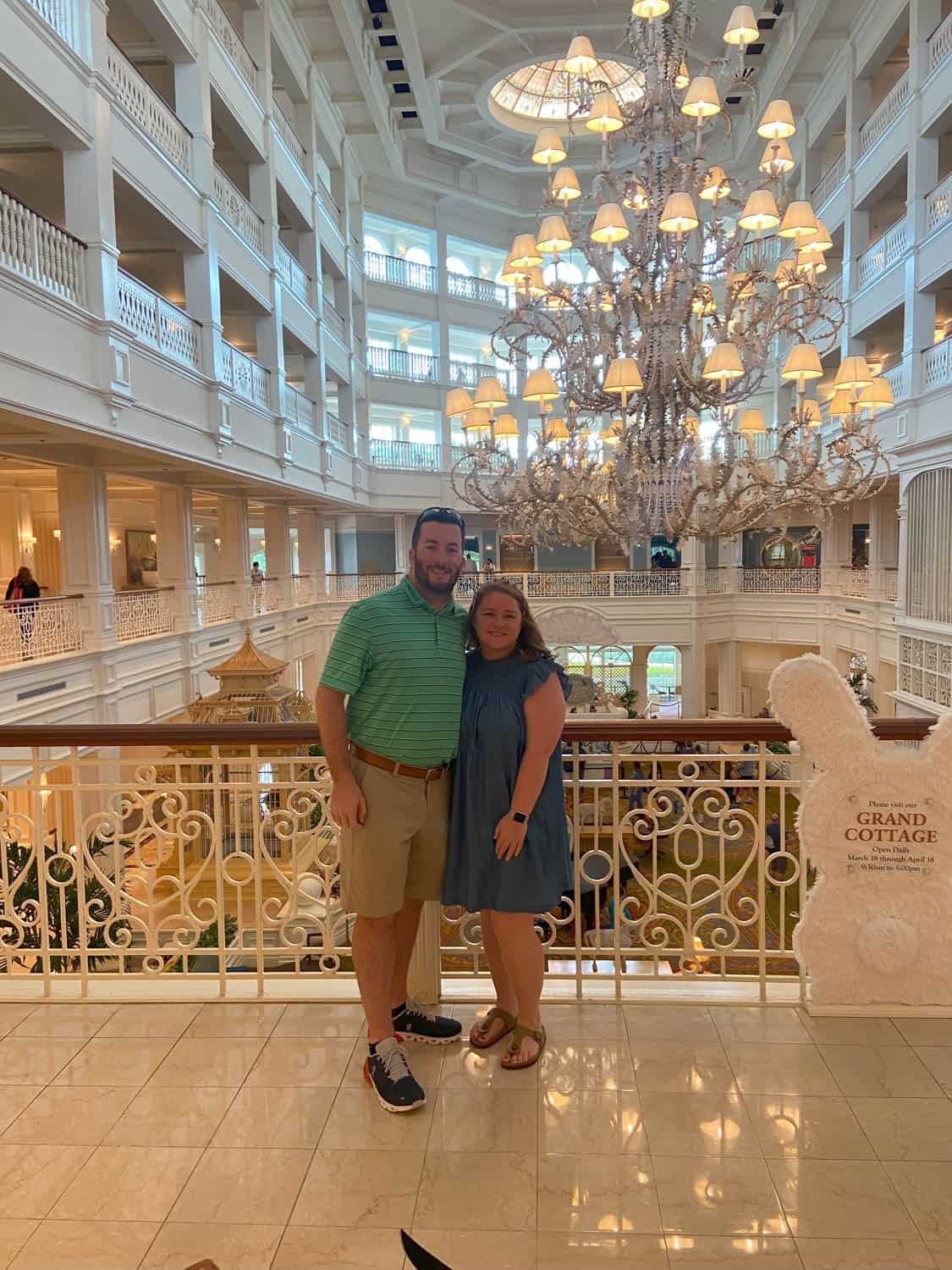Olivia-Williams-05 A man and woman stand together inside a large, elegant hotel lobby with white railings, marble floors, and a grand chandelier overhead.