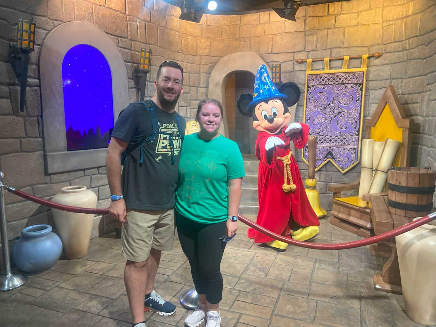 Olivia-Williams-06 Two people smile in front of a rope barrier with Sorcerer Mickey Mouse in a medieval-themed room with stone walls and decor.