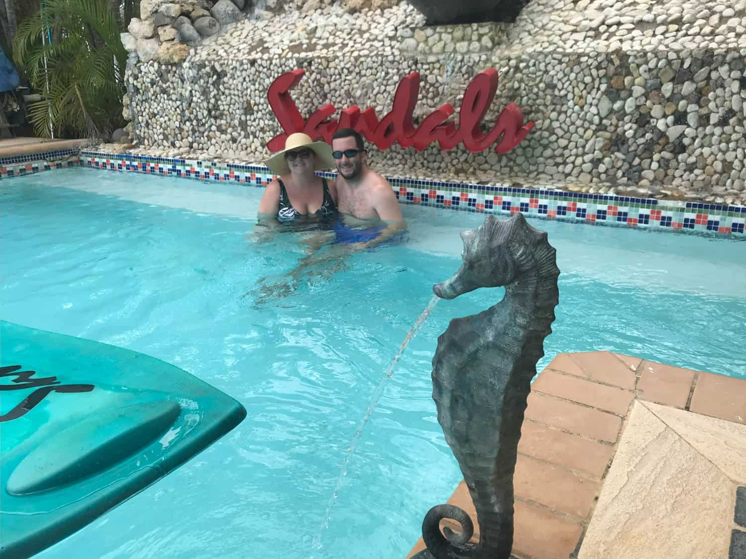 Olivia-Williams-07 Two people pose together in a pool near a Sandals sign, with a seahorse fountain statue in the foreground.