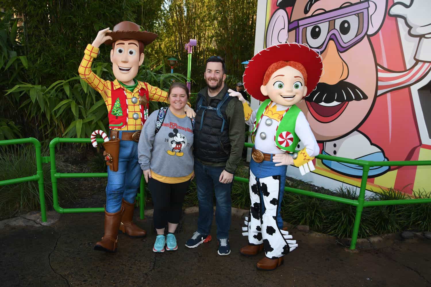 Olivia-Williams-08 Two people stand between Woody and Jessie costumed characters at a theme park, with a large Mr. Potato Head image in the background.