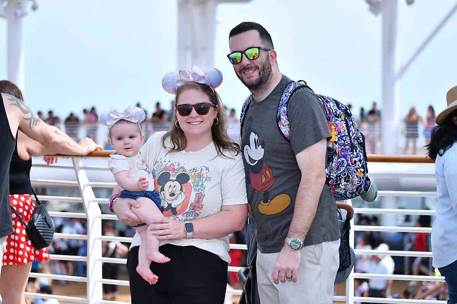 Olivia-Williams-09 A woman holding a baby and a man stand together on a ship deck, all wearing Disney-themed clothing and accessories, with people and railings in the background.