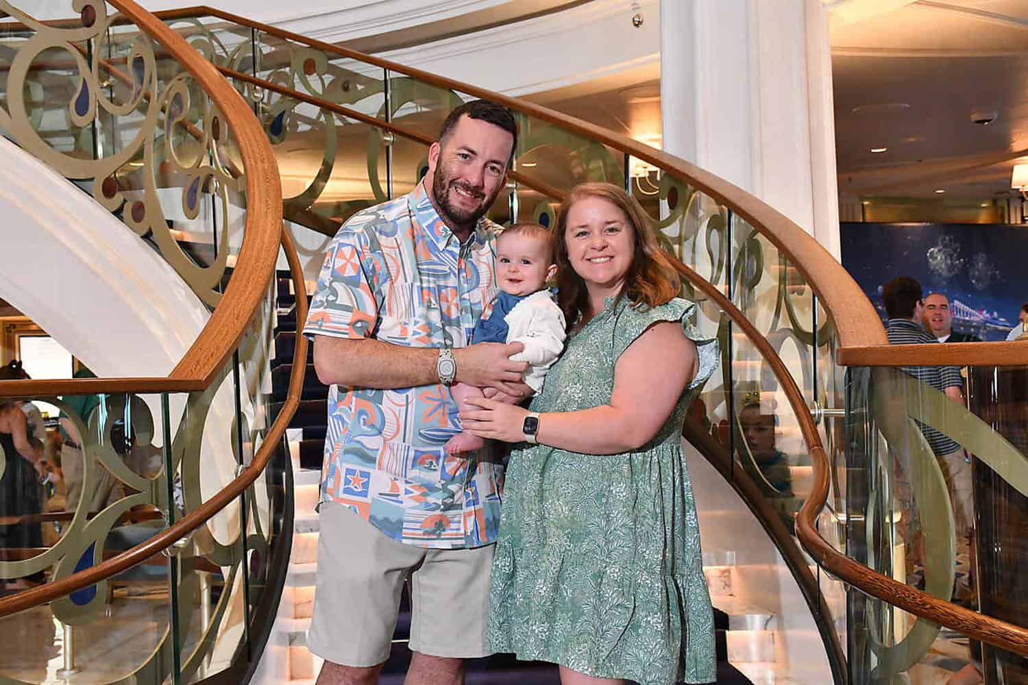 Olivia-Williams-10 A man, woman, and baby stand together smiling in front of a decorative spiral staircase in a well-lit indoor setting.