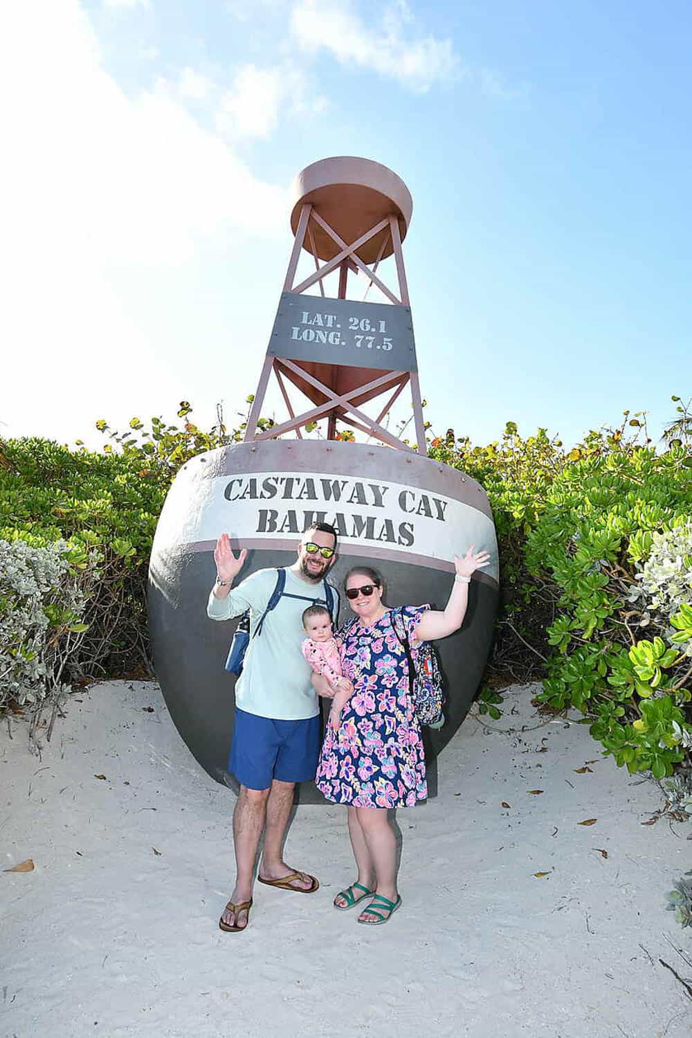 Olivia-Williams-11 A family of three stands in front of a large Castaway Cay Bahamas buoy, smiling and waving at the camera on a sandy beach.