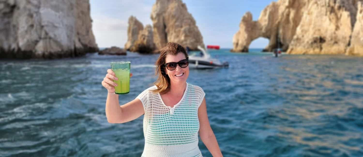 Anna-Swenson-201 Woman in sunglasses holds a green drink on a boat, with rocky sea cliffs and an arch formation in the background.