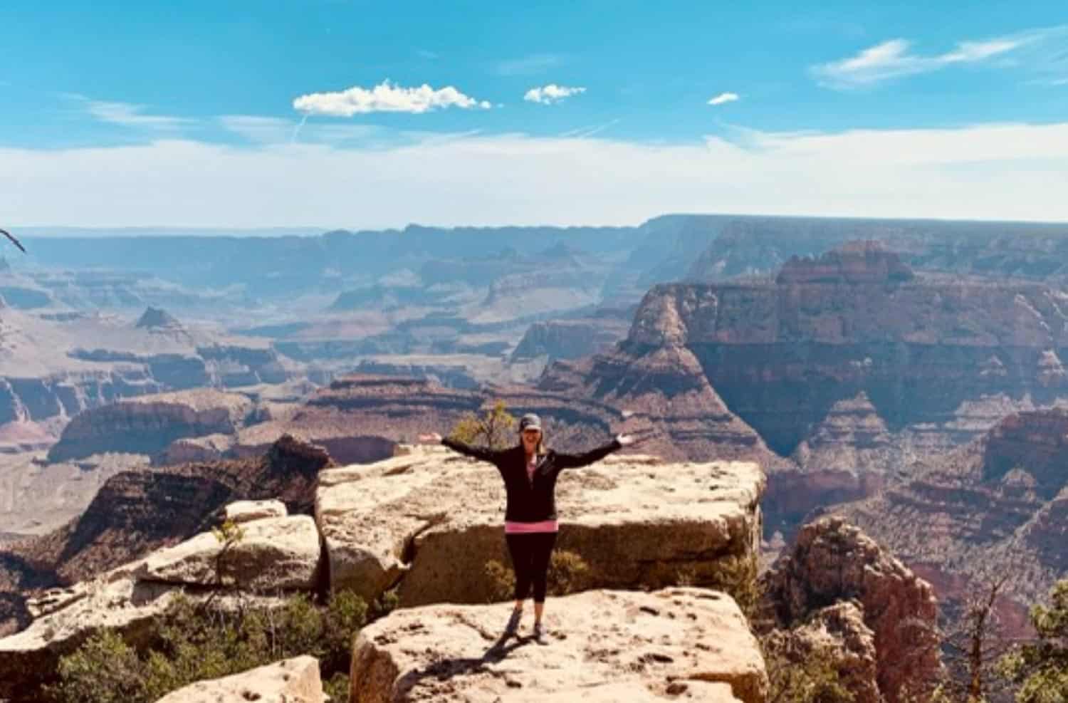 Anna-Swenson-202 A person stands with arms outstretched on a rocky ledge overlooking the expansive landscape of the Grand Canyon under a sunny sky.