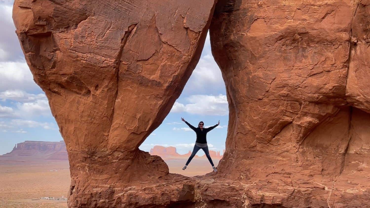 Anna-Swenson-203 A person stands with arms and legs outstretched between two large red rock formations in a desert landscape.