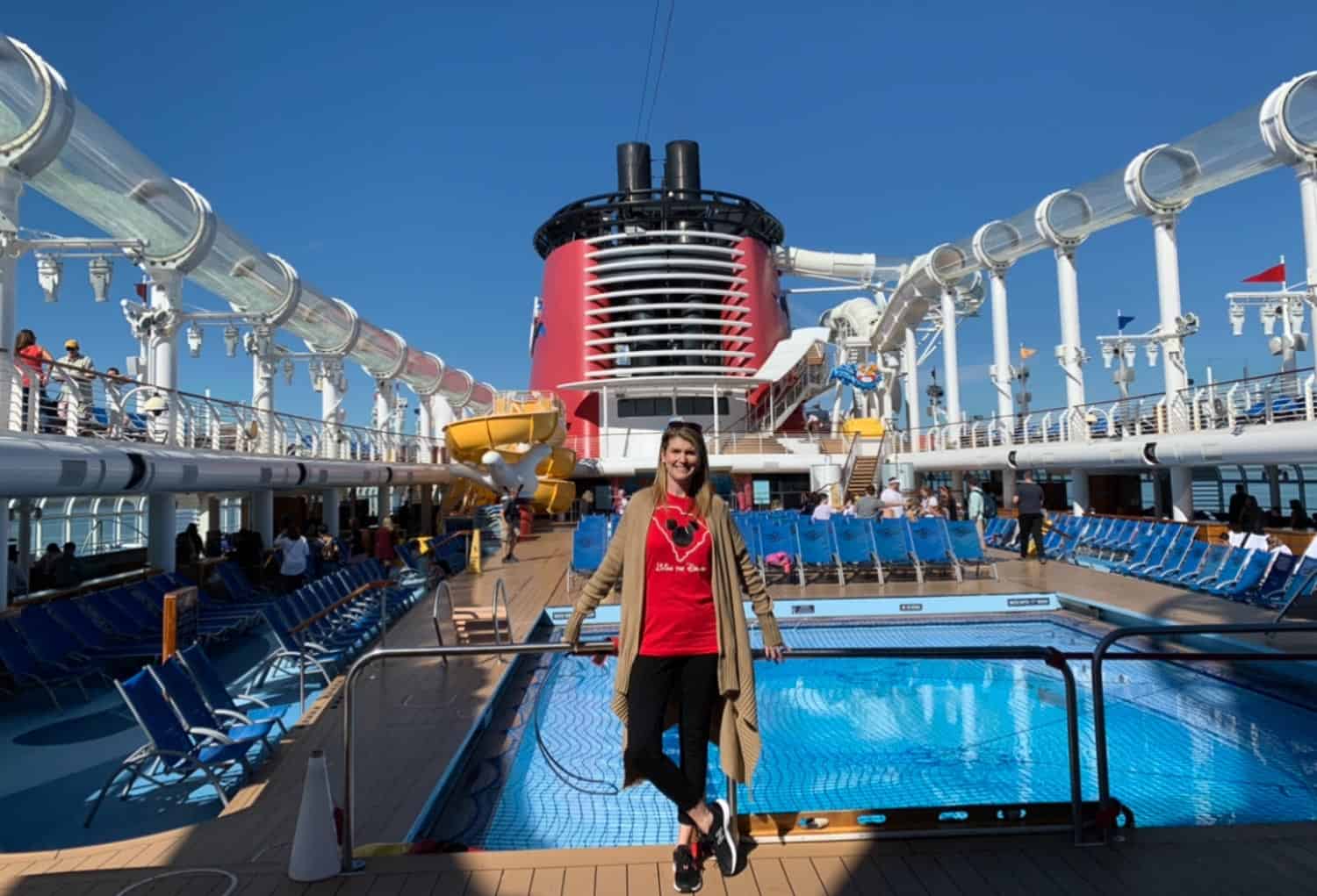 Anna-Swenson-204 A woman stands by an empty pool on the deck of a cruise ship, with waterslides and lounge chairs visible under a clear blue sky.