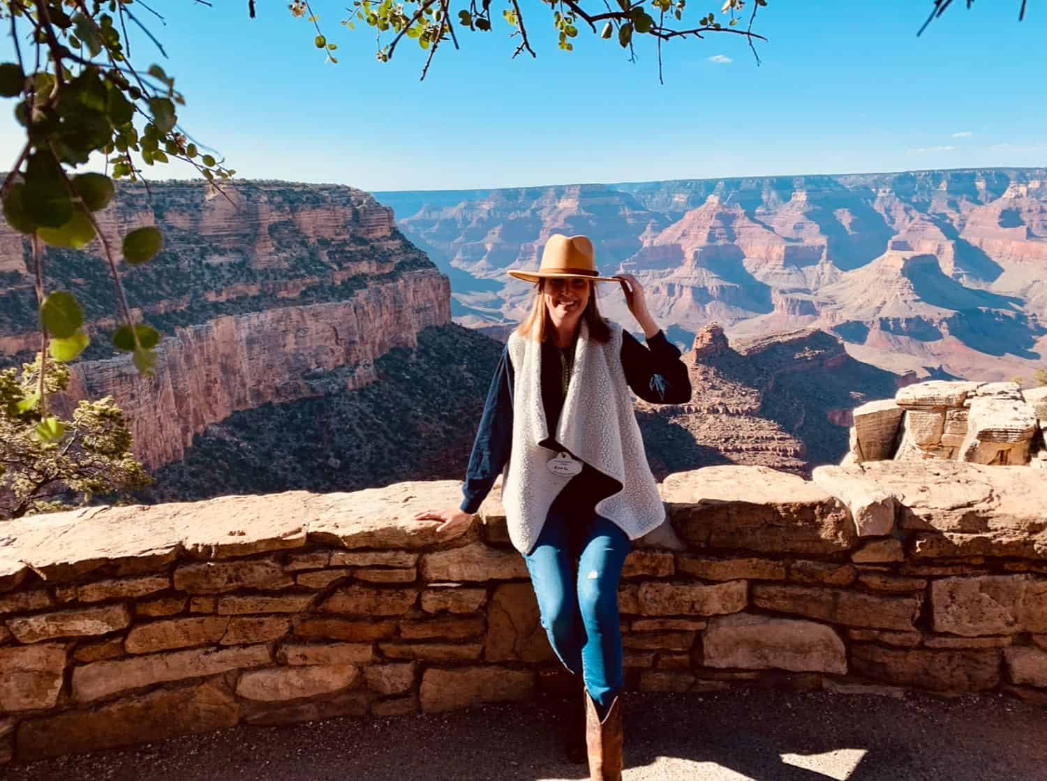 Anna-Swenson-206 Person wearing a hat and vest leans against a stone wall with the Grand Canyon landscape in the background on a sunny day.