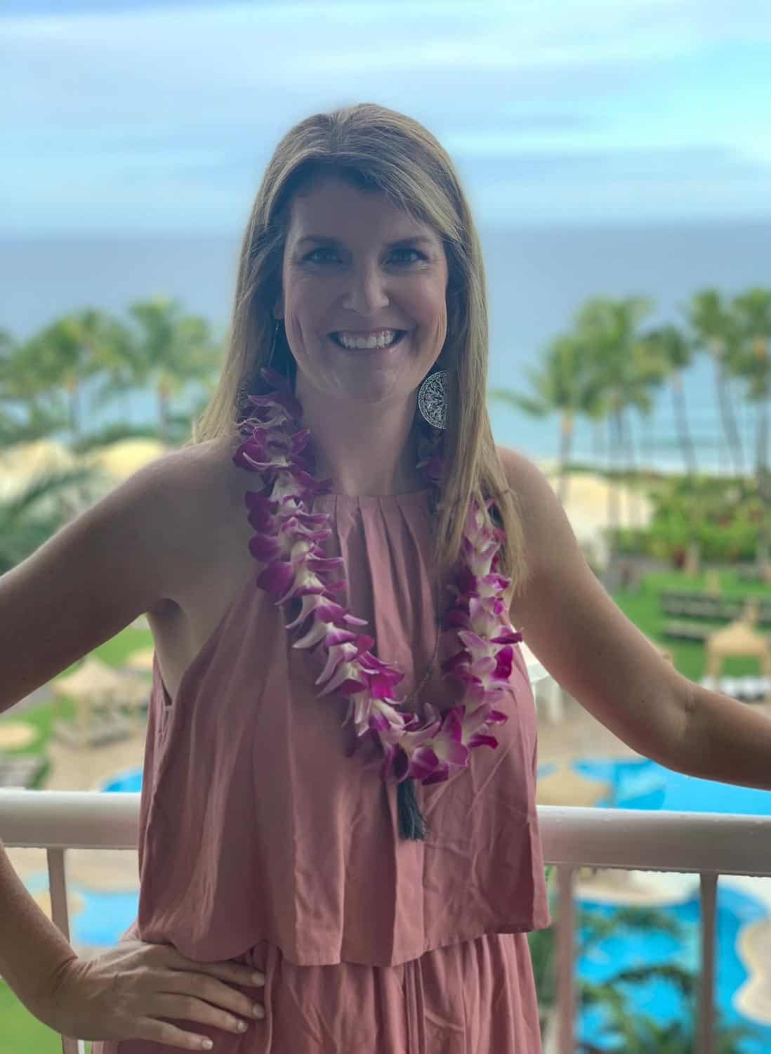 Anna-Swenson-207 Woman in a pink sleeveless dress wearing a purple lei stands on a balcony with palm trees, ocean, and pool visible in the background.