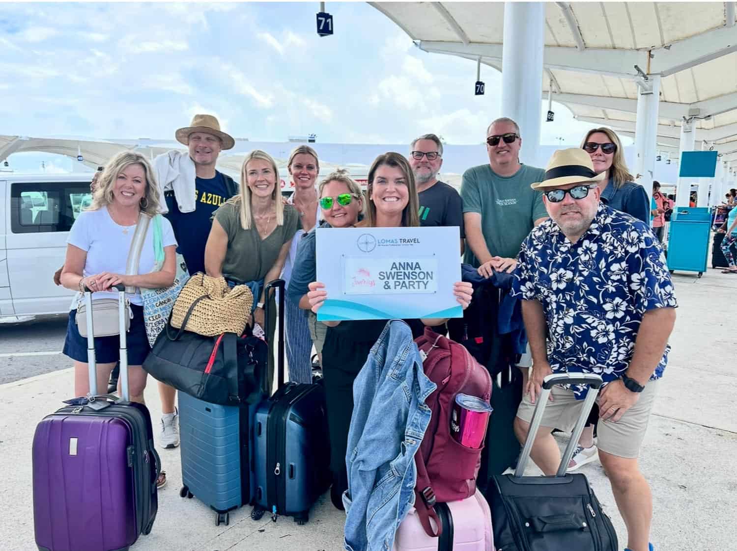 Anna-Swenson-208 A group of ten adults with luggage pose outdoors at an airport; one person holds a sign that reads Anna Swenson and Party.
